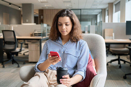 Hispanic latin young woman student holding smartphone and coffee sitting in modern space studying on cell, browsing, checking apps, texting using mobile phone in university campus or creative office.