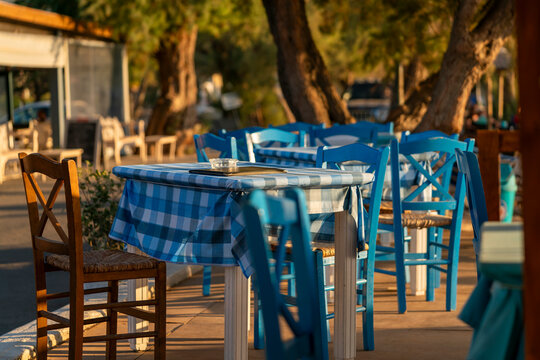 Empty seaside terrace on Lesbos with blue chairs and tables in warm evening sunlight, peaceful Mediterranean dining setting under trees with soft golden light atmosphere
