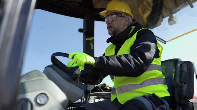 Close-up of a focused male construction worker in safety gear operating a heavy machinery vehicle, steering and controlling the equipment at a sunny building plot.