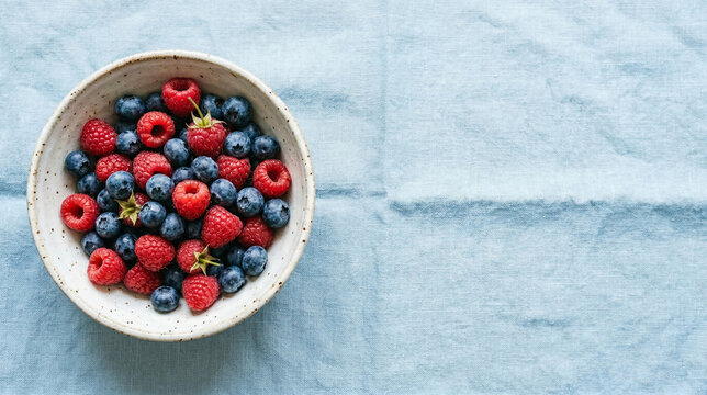 Flat lay of fresh blueberries and raspberries in a rustic ceramic bowl on a light blue linen background with copy space for text
