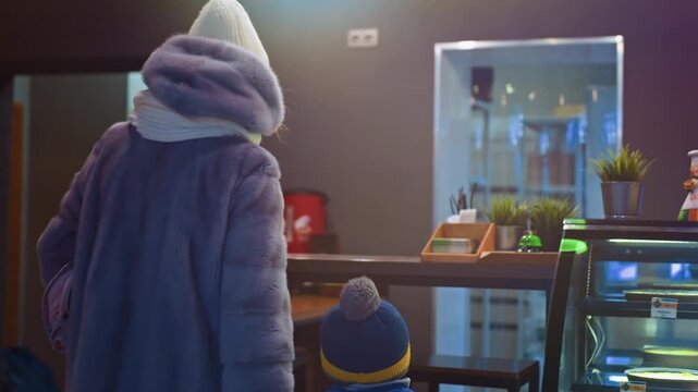 Mom and son at pastry counter inside warm cafe, woman in gray coat and knit scarf stands with child in blue jacket and pompom beanie looking at illuminated glass display of cakes and desserts, soft