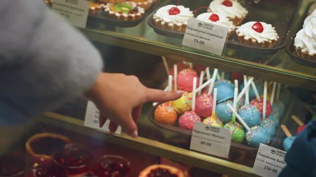 Mom son at bakery display pointing, mother finger hovering over colorful cake pops behind glass, child in blue jacket and beanie watching eagerly, rows of tarts and cupcakes on shelves, warm cozy