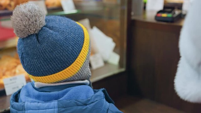 Mom and son at bakery counter choosing treat, warm gesture as adult hand guides child in blue beanie with pompom, glass case filled with croissants and rolls, price tags and labels visible, cozy