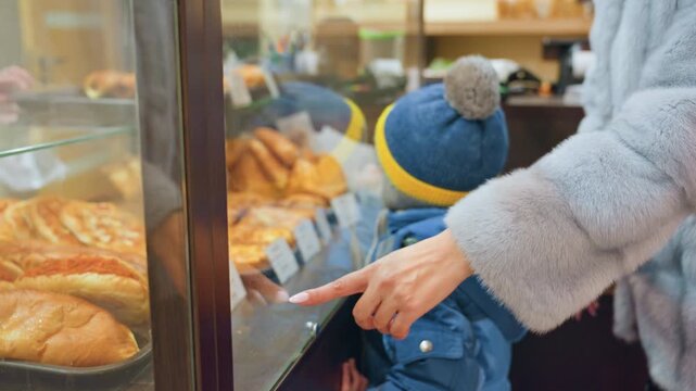 Mom and son at bakery display, mom in gray coat pointing at pastries through glass case as young son in blue hat watches, croissants and baguettes on trays, cozy winter shopping moment, mother guiding