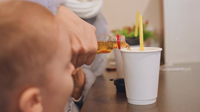 White mom and son pouring syrup into paper cup, closeup of nurturing hand and curious toddler watching, yellow straw, cozy indoor table, soft scarf and warm tones evoke calm morning bonding