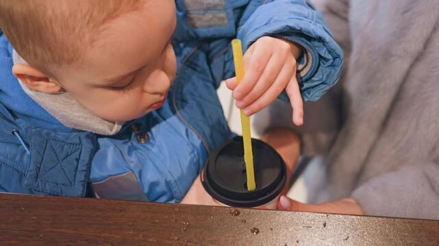 Caucasian mom assists toddler son sipping from travel cup with yellow straw at wooden counter, blue jacket, gentle hand guidance, warm indoor lighting, close nurturing moment, focused expression,