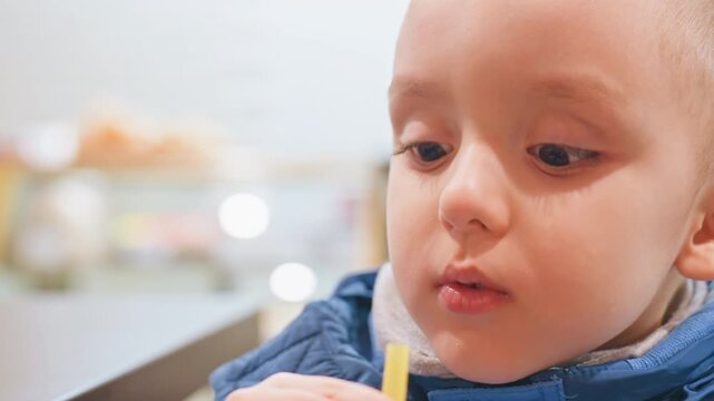 Toddler son sipping straw in cafe, closeup portrait capturing curious expression and puckered lips as off camera mom offers warm cup, soft bokeh background, cozy lighting, blue jacket, candid family