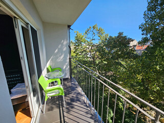 Naklejka premium Balcony with a green chair and a table. The table has a glass top. The balcony is overlooking a tree-lined street