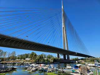 Obraz premium Bridge spans a river with a large number of boats docked on the water. The bridge is a long, thin structure with a clear blue sky above it