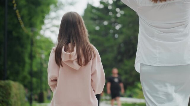 Mom daughter walking past runner in park, rear closeup of small girl in pink hoodie and mother in white blouse, passing active jogger on treelined trail, casual pace, urban park atmosphere, soft