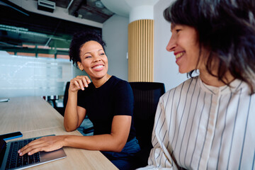 Two diverse business women laughing and discussing work during an office meeting, showing teamwork and positive corporate communication