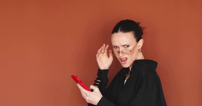 Young woman turning head with WTF face to camera lowering glasses and looking with reproach, using smartphone and feeling annoyed by news, stylish female model in black shirt against orange background