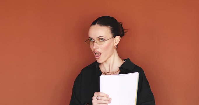 Shocked stylish woman looking at documents with open mouth and surprised expression on brown background. Young female office worker or student holding paper folder and feeling amazed or stressed