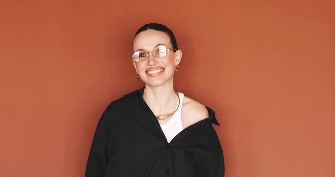 Excited stylish woman pointing down with both hands and laughing joyfully against brown studio background. Young female model in rimless glasses and black shirt showing empty space for text or link