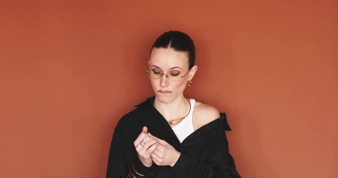 Bored young woman looking aside and nervously fidgeting with fingers or fingernails, adjusting rings and glasses on orange background, annoyed or impatient female model waiting and posing to camera