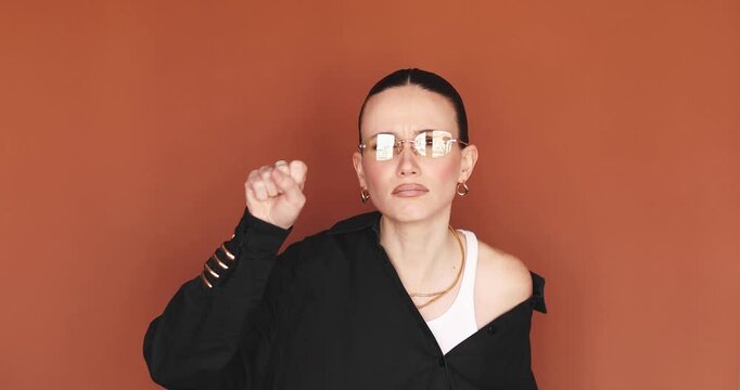 Confident young woman in yellow glasses squinting to look closer and making a fist to knock or threaten, standing with aggressive expression against terracotta background wall