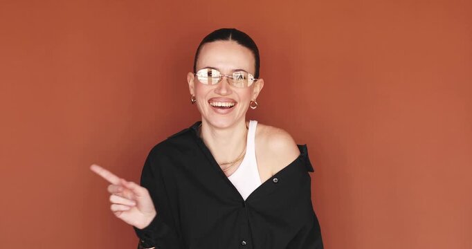Playful young woman in yellow glasses pointing to herself asking "Me?" and then laughing, saying "No" and making negation finger wag gesture against terracotta background wall