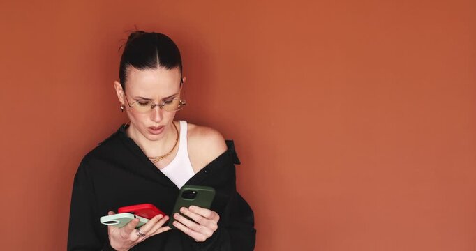 Shocked woman looking at three smartphones simultaneously with confused face expression. Surprised girl checking notifications on multiple mobile devices on terracotta background, information shock
