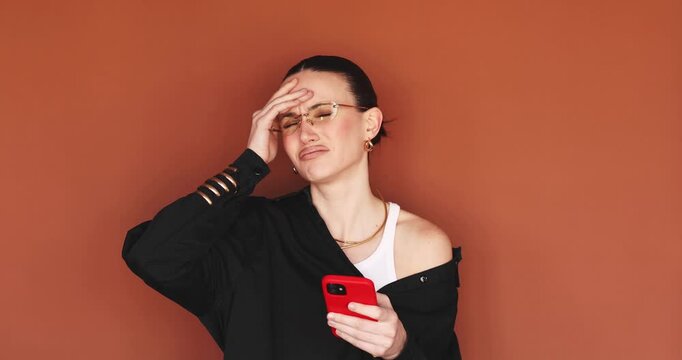 Disappointed young woman looking at smartphone and doing facepalm gesture. Upset girl reading bad news on phone, feeling frustrated and annoyed on terracotta background, negative online experience
