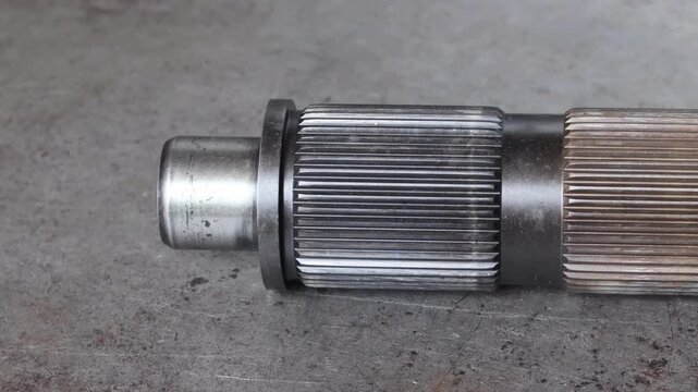 Panning shot of a heavy-duty steel spline shaft gear on a metal workshop floor, showing industrial texture and mechanical details of the drivetrain component.
