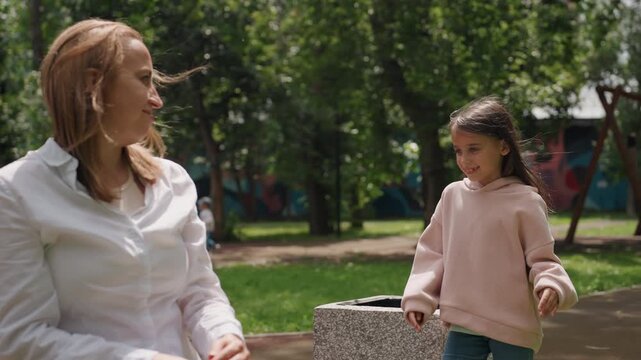 Park bench caucasian mom comforts daughter with hug and gentle wipe sunny green park with trees and distant playground mother sits on stone bench while daughter approaches in pink hoodie interaction