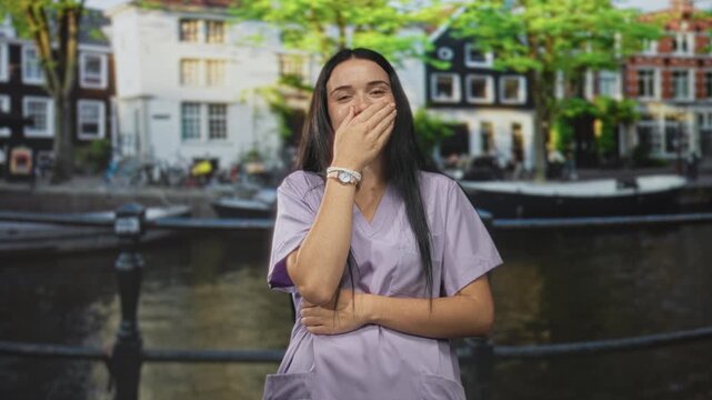 Woman nurse in purple scrubs laughs while covering mouth with right hand beside canal railing and historic building, smiling eyes visible; care compassion joy.