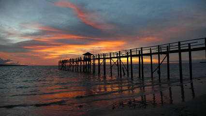 Obraz premium Golden and pink hues paint the sky as the sun sets behind the tranquil pier at the beach, reflecting in calm waters