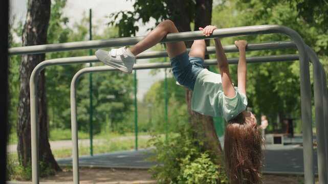 White daughter hanging across park bars, middistance shot of athletic girl performing inverted hang on metal rails, curly hair falling, sunny urban green space and playful strength.