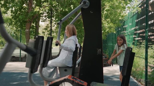 White mom and daughter during outdoor workout, mother pulls handles while daughter watches and helps from behind, energetic encouragement, leafy park setting, casual athletic vibe