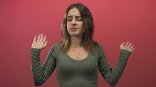 Young hispanic woman with heart necklace raises both hands in a shrug gesture in a pink studio; resignation.