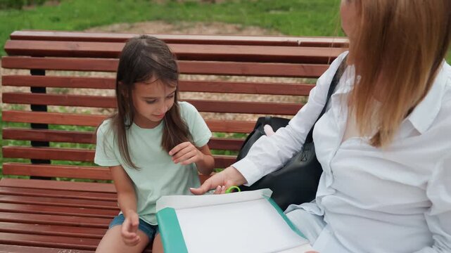 Mom and daughter drawing together on bench with sketchbook and crayons, mother guiding childs hand across page, focused and playful outdoor lesson, gentle interaction and shared creativity