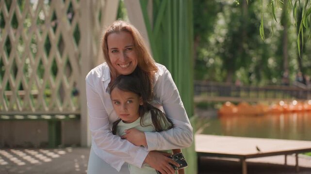 Mom and daughter hugging under willow tree, mother leaning over child with warm smile, shaded gazebo setting by calm water, soft dappled light, intimate protective moment and playful calm in summer
