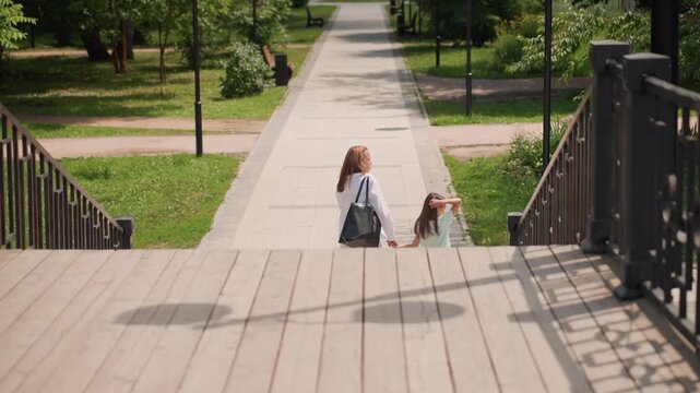 Mom and daughter descending wooden steps, mom carrying tote bag, daughter skipping near railing, sunlit park path, playful pause, dramatic lamp shadow on deck, candid movement, summer mood, casual