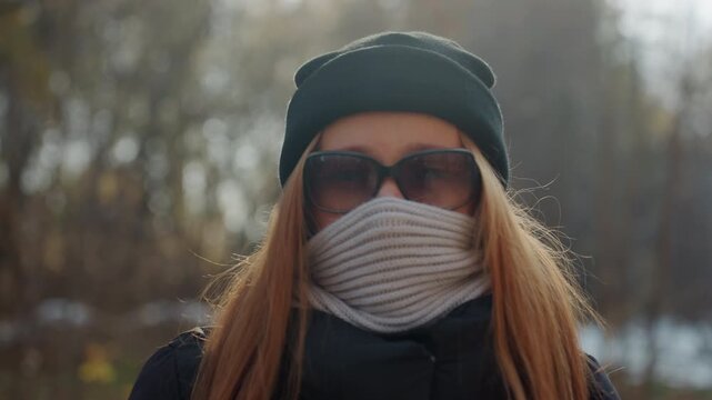 Caucasian woman masked in park, red hair peeking from knit hat and scarf covering lower face, sunglasses obscuring eyes, black beanie and backpack, puffy jacket walking along woodland trail, turns