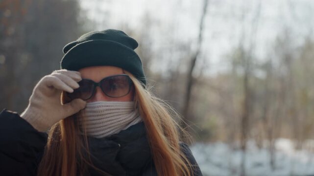 White woman unmasking in snowdusted forest, lifting sunglasses with gloved hand to reveal eyes, scarf and beanie, soft sunlight and backlight, expression shifts from guarded to relieved, traveler