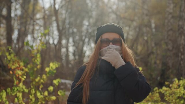 Red haired white woman walking alone in autumn woods, wearing black coat, knit beanie, scarf and gloves, backpack on shoulders, sunglasses on face, cautious gaze scanning surroundings, tense mood