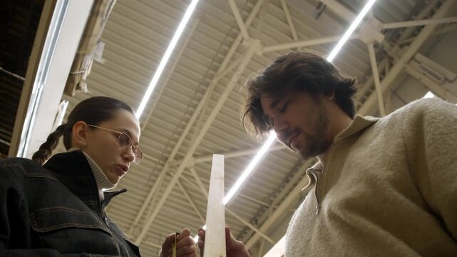Shot shows young couple measuring wooden batten length in hardware store, viewed from shopping cart perspective with bright overhead lights and shelves in background.