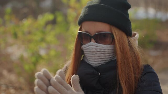 Gloved white woman adjusts hands in forest closeup, knitted scarf and sunglasses conceal face, focused movements convey preparation and intent of a snatcher in chilly sunlit woodland