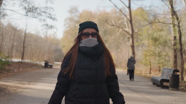 Caucasian woman walking in park, bundled in puffer jacket and beanie, scarf covering lower face, steady stride along leafstrewn path with birch trees and benches, distant pedestrians and empty benches