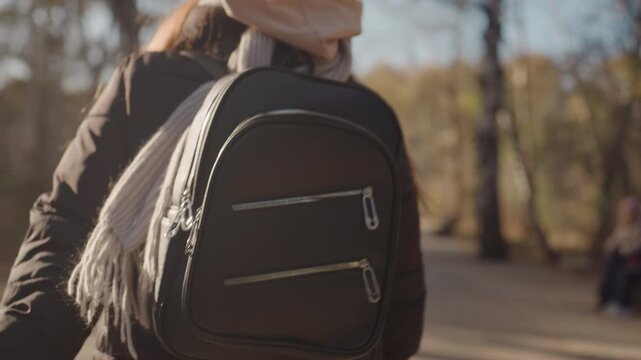 Woman with backpack walking in park. Autumn afternoon, puffer jacket and scarf, closeup on bag and straps, subtle tension suggests possible snatcher nearby, cinematic warm tones and candid street
