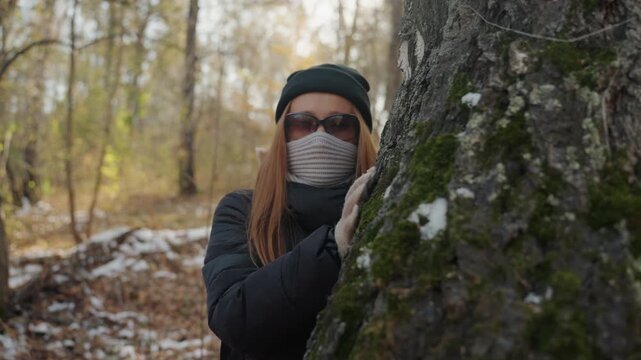 Woman prowler hides behind mossy tree, peeking around trunk in snowy autumn woodland, sunglasses and knit scarf conceal face, beanie and padded jacket signal cold, slow cinematic pan builds suspense