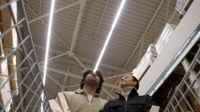Couple pushes shopping cart forward browsing goods in hardware store from cart view. They stroll aisle studying shelves under high ceiling.