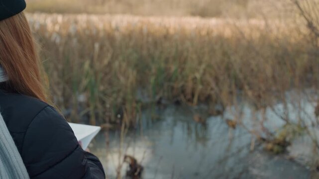 Redhead woman holding map by marsh in autumn light, coat and beanie, standing at waters edge amid tall reeds and puddle reflections quiet, misty horizon and muted colors create contemplative mood