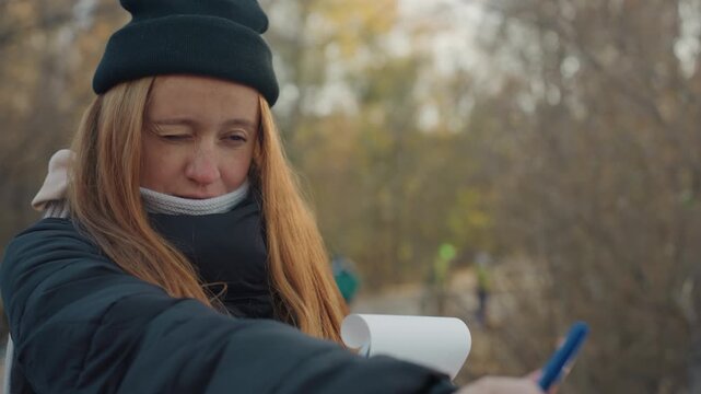 Woman taking notes in autumn park, bundled in coat and beanie, focused expression while flipping pages and jotting observations, background of bare trees and scattered leaves, cool light, candid
