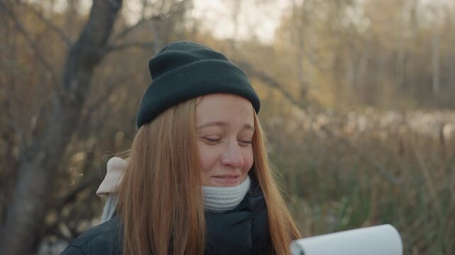 Caucasian woman hiding notebook outdoors, playful snatcher vibe, smiling behind notepad among autumn reeds, beanie and knit scarf, candid closeup showing smirk and gentle gaze, warm golden light,
