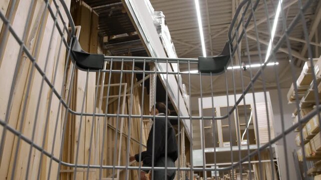 POV shot from shopping cart shows man pushing trolley toward girlfriend at wooden boards rack in large hardware store, both preparing to select lumber together.