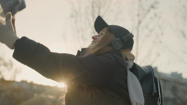 Caucasian redhead woman holding stuffed rabbit near sunset in urban park, winter coat, cap and headphones, backpack slung over shoulder, gloved hands cradle plush toy, joyful smile and soft laugh,