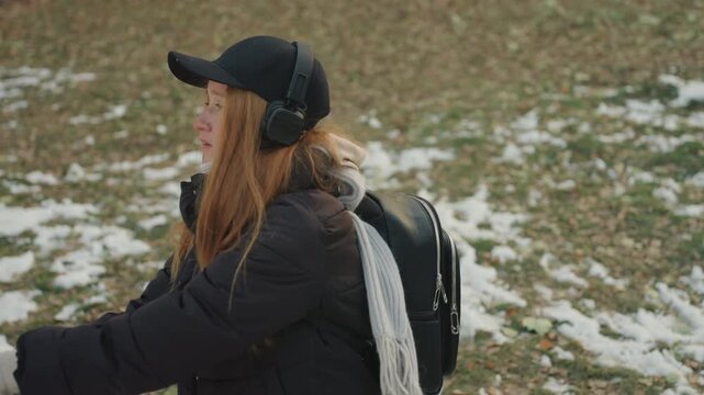 Park scene white redhead woman retrieves plush rabbit with gloved hands after snatcher incident light snow patches on grass, backpack, puffer coat and cap, reaching arms, hopeful smile, candid