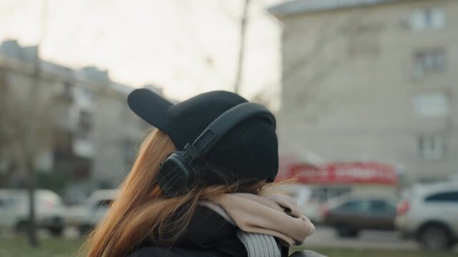 Woman wearing headphones and cap turns toward possible prowler near urban street, red hair visible under beanie, scarf and coat, clutching small plush toy, cars and apartment buildings blurred