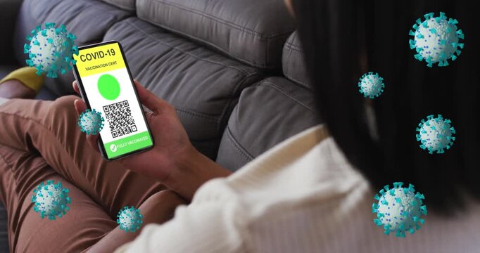 Animation of covid 19 cells over woman relaxing at home with digital covid passport on phone screen